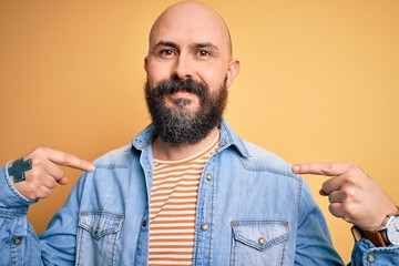 Handsome bald man with beard wearing casual denim jacket and striped t-shirt looking confident with...