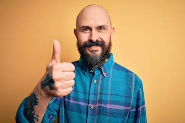 Naklejka premium Handsome bald man with beard wearing casual shirt standing over isolated yellow background doing happy thumbs up gesture with hand. Approving expression looking at the camera showing success.