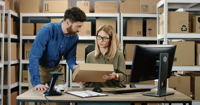 Caucasian woman postal worker working at computer in office store. Postman bringing carton box and female colleague redistering it while couple talking. Mail delivery registration.