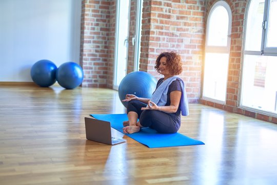 Middle age beautiful sportswoman smilig happy. Sitting on mat with smile on face using laptop at home
