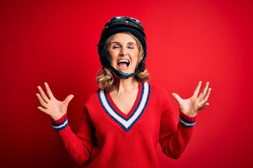 Middle age beautiful blonde motorcyclist woman wearing moto helmet over red background crazy and mad shouting and yelling with aggressive expression and arms raised. Frustration concept.