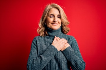 Middle age beautiful blonde woman wearing casual turtleneck sweater over red background smiling with hands on chest with closed eyes and grateful gesture on face. Health concept.