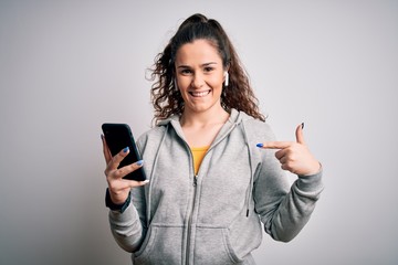 Beautiful sportswoman with curly hair listening to music using smartphone and earphones with surprise face pointing finger to himself