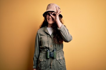 Young beautiful tourist woman on vacation wearing explorer hat and binoculars doing ok gesture with hand smiling, eye looking through fingers with happy face.