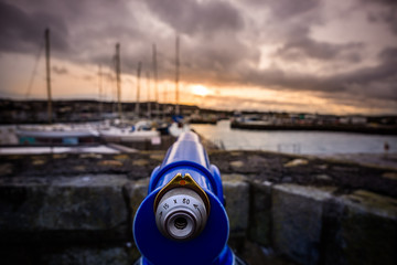 Selective focus on blue telescope at Howth harbour with blurred yachts and sunset in background, Dublin, Ireland