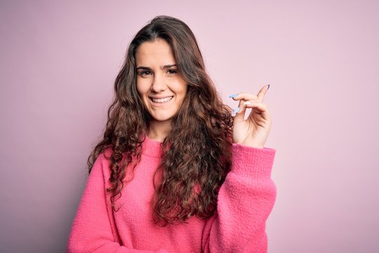 Young beautiful woman with curly hair wearing casual sweater over isolated pink background with a big smile on face, pointing with hand and finger to the side looking at the camera.