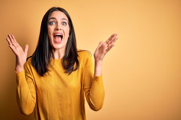 Young brunette woman with blue eyes wearing casual sweater over yellow background celebrating crazy and amazed for success with arms raised and open eyes screaming excited. Winner concept