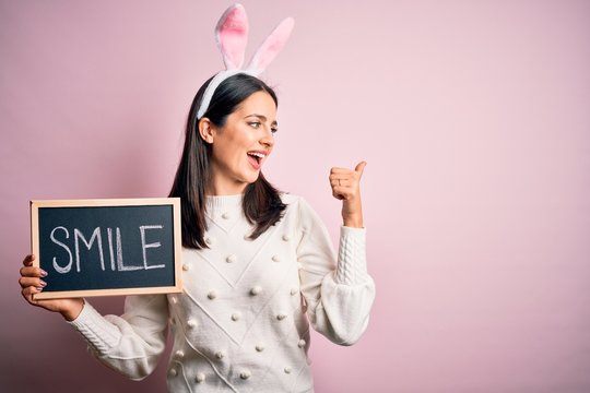 Young caucasian woman wearing easter rabbit ears and holding blackboard with smile word pointing and showing with thumb up to the side with happy face smiling