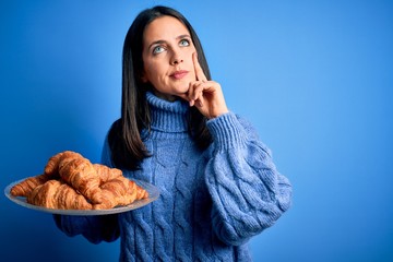 Young woman with blue eyes holding sweet croissants for breakfast over isolated background serious...