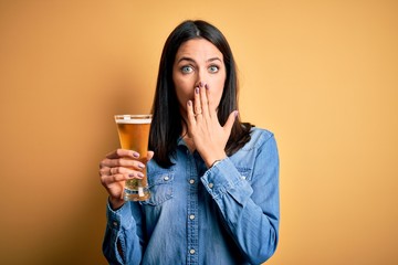 Young woman with blue eyes drinking glass of beer standing over isolated yellow background cover mouth with hand shocked with shame for mistake, expression of fear, scared in silence, secret concept