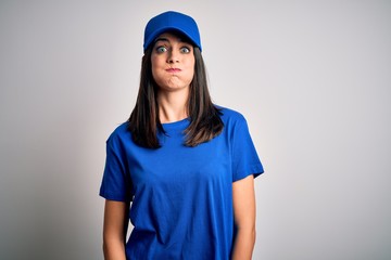 Young delivery woman with blue eyes wearing cap standing over blue background puffing cheeks with funny face. Mouth inflated with air, crazy expression.