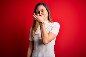 Beautiful blonde woman with blue eyes wearing casual white t-shirt over red background smelling...