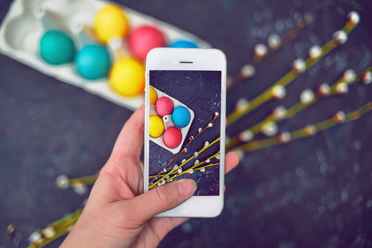 Happy Easter. Painted colored, pastel eggs in an egg carton. Flowering willow fur seal, flowers on an abstract background of dark color. The phone takes a photo of the holiday, the girl takes a photo