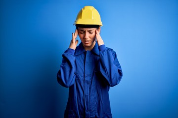 Young beautiful worker woman with blue eyes wearing security helmet and uniform with hand on head for pain in head because stress. Suffering migraine.