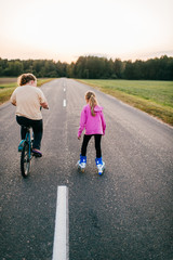 Fototapeta premium Pretty young girl in pink hoody roller-skates on the village road in the evening with her elder sister