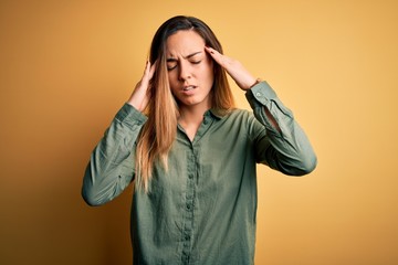 Young beautiful blonde woman with blue eyes wearing green shirt over yellow background with hand on head for pain in head because stress. Suffering migraine.