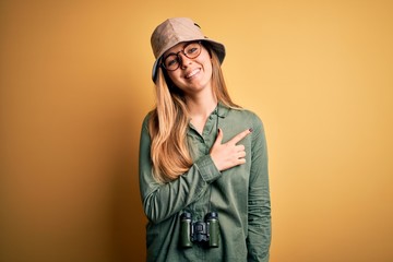 Beautiful blonde explorer woman with blue eyes wearing hat and glasses using binoculars cheerful with a smile of face pointing with hand and finger up to the side with happy and natural expression 