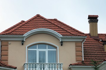Obraz premium brown attic of a private boom with a large window and a white balcony under a red tiled roof with a brick chimney against the sky