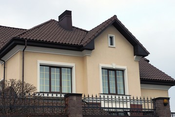 brown private house with windows under a tiled roof against a gray sky behind a fence 