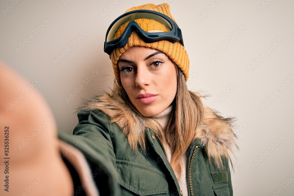 Poster Young brunette skier woman wearing snow clothes and ski goggles make selfie by camera with a confident expression on smart face thinking serious