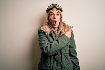 Young brunette skier woman wearing snow clothes and ski goggles over white background Surprised pointing with finger to the side, open mouth amazed expression.