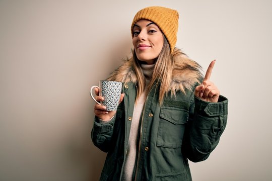 Young beautiful brunette woman wearing snow clothes drinking mug of coffee surprised with an idea or question pointing finger with happy face, number one