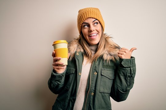Young beautiful brunette woman wearing snow clothes drinking cup of coffee pointing and showing with thumb up to the side with happy face smiling