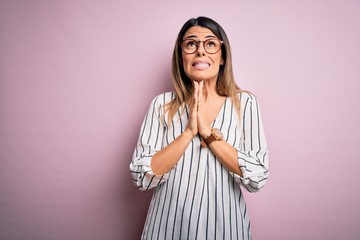 Young beautiful woman wearing casual striped t-shirt and glasses over pink background begging and...