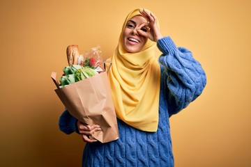 Young beautiful brunette arab woman wearing islamic hijab holding paper bag with food with happy...