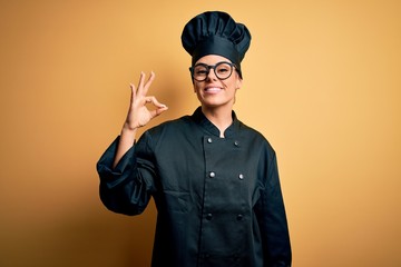 Young beautiful brunette chef woman wearing cooker uniform and hat over yellow background smiling positive doing ok sign with hand and fingers. Successful expression.