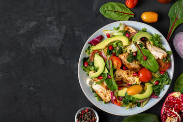 Grilled chicken breast and avocado salad with mixed greens, tomatoes and pomegranate in a plate on dark background