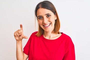 Beautiful redhead woman wearing casual red t-shirt over isolated background smiling and confident gesturing with hand doing small size sign with fingers looking and the camera. Measure concept.
