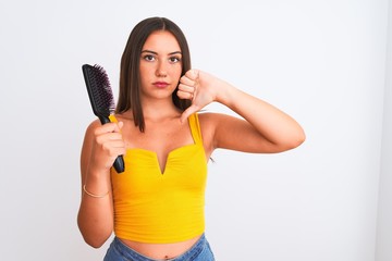 Young beautiful girl holding hair comb standing over isolated white background with angry face,...