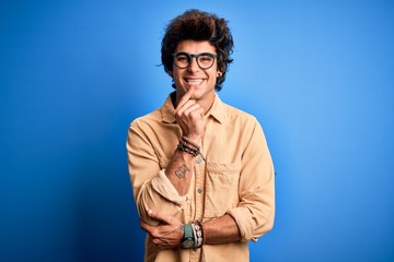 Young handsome man wearing casual shirt standing over isolated blue background looking confident at the camera with smile with crossed arms and hand raised on chin. Thinking positive.