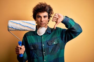 Young handsome painter man holding roller standing over isolated yellow background with angry face, negative sign showing dislike with thumbs down, rejection concept