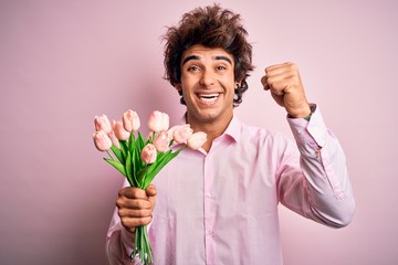 Young handsome man holding flowers standing over isolated pink background screaming proud and celebrating victory and success very excited, cheering emotion