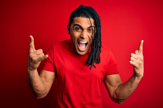 Young handsome african american afro man with dreadlocks wearing red casual t-shirt shouting with crazy expression doing rock symbol with hands up. Music star. Heavy music concept.