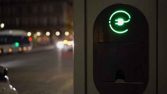 Electric vehicle charging station close up. Night city lights, facades of beautiful buildings and a passing by cars in the background. Refueling, charging a car in the parking lot. Ultra HD footage