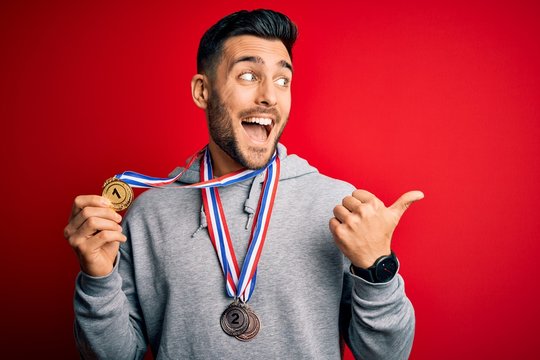 Young handsome succesful man winning medals standing over isolated red background pointing and showing with thumb up to the side with happy face smiling