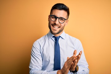 Young handsome businessman wearing tie and glasses standing over yellow background clapping and applauding happy and joyful, smiling proud hands together