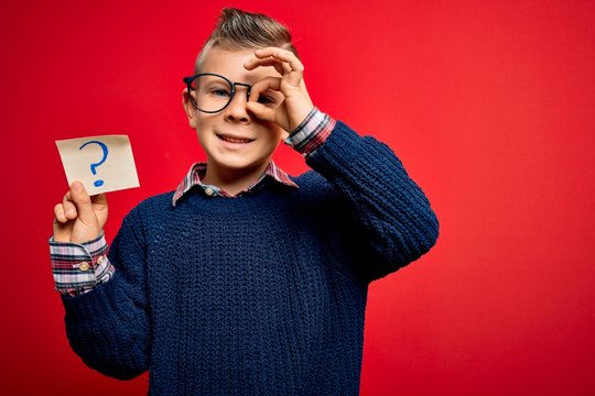 Young little caucasian kid wearing glasses holding paper note with question mark sign with happy face smiling doing ok sign with hand on eye looking through fingers