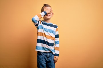 Young little caucasian kid with blue eyes wearing colorful striped shirt over yellow background smiling and laughing with hand on face covering eyes for surprise. Blind concept.