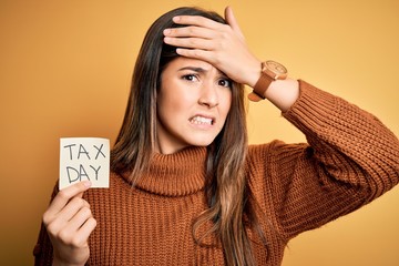 Young beautiful girl holding reminder paper with tax day message over yellow background stressed...