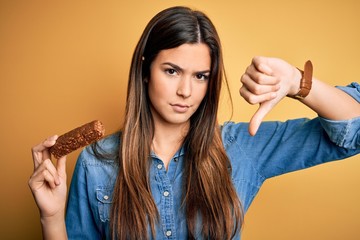 Young beautiful girl holding healthy protein bar standing over isolated yellow background with...