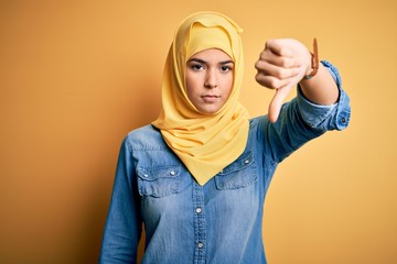 Young beautiful girl wearing muslim hijab standing over isolated yellow background looking unhappy and angry showing rejection and negative with thumbs down gesture. Bad expression.