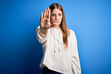 Young beautiful redhead sporty woman wearing sweatshirt over isolated blue background doing stop...