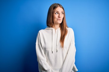 Young beautiful redhead sporty woman wearing sweatshirt over isolated blue background smiling looking to the side and staring away thinking.