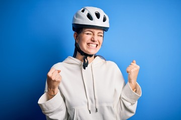 Young beautiful redhead cyclist woman wearing bike helmet over isolated blue background very happy and excited doing winner gesture with arms raised, smiling and screaming for success. Celebration