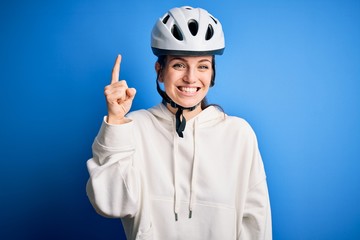 Young beautiful redhead cyclist woman wearing bike helmet over isolated blue background pointing finger up with successful idea. Exited and happy. Number one.