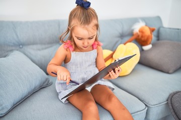 Young beautiful blonde girl kid enjoying play school with toys at kindergarten, smiling happy drawing on clipboard